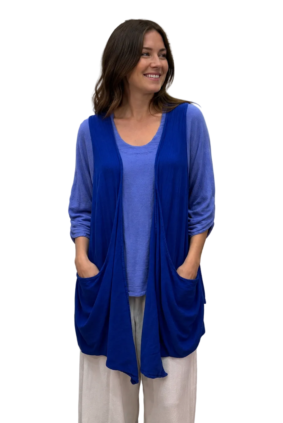 A woman modeling a vibrant royal blue open-front cardigan with pockets over a periwinkle blue top and cream pants, standing in front of a white fabric backdrop.