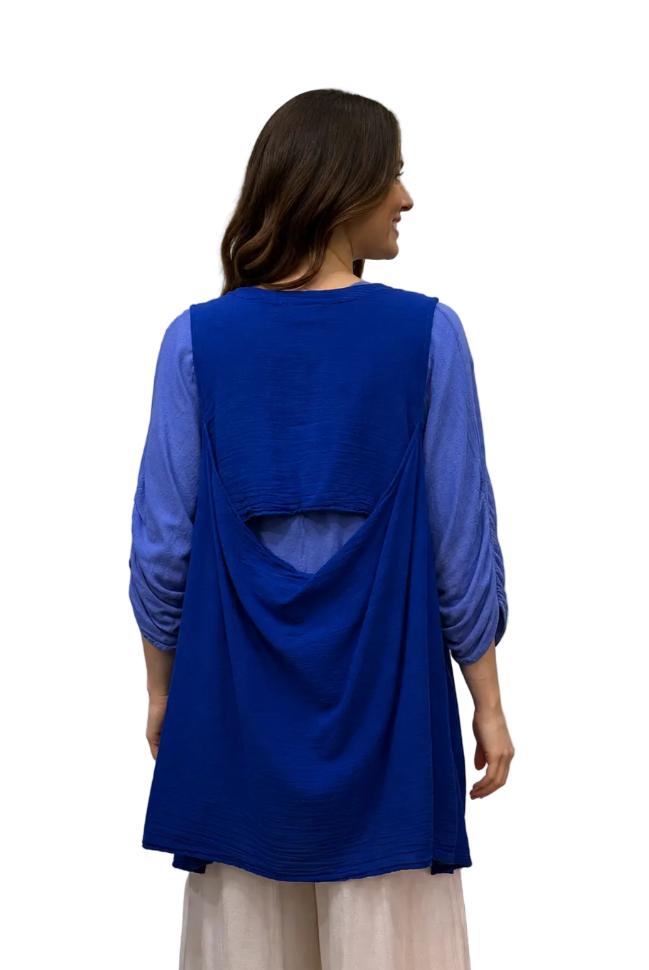 A woman viewed from the back wearing a bright royal blue sleeveless tunic layered over sheer light blue three-quarter sleeves, paired with light cream wide-leg pants, standing in front of a white fabric backdrop.