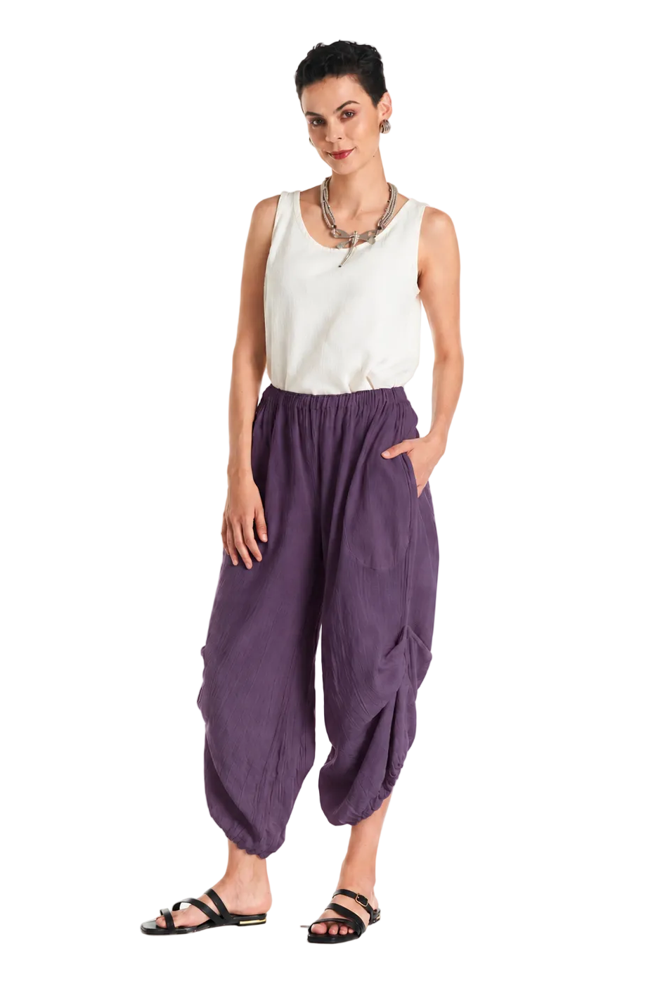 A woman modeling loose, gathered harem-style pants in a muted purple color, paired with a white sleeveless top and black sandals, against a plain white studio background.