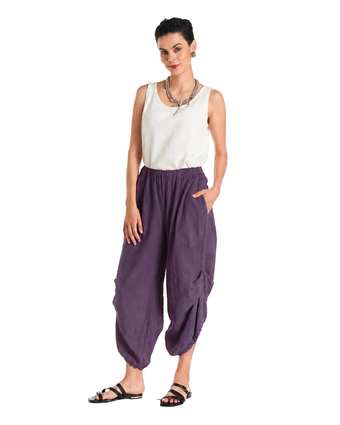 A woman modeling loose, gathered harem-style pants in a muted purple color, paired with a white sleeveless top and black sandals, against a plain white studio background.