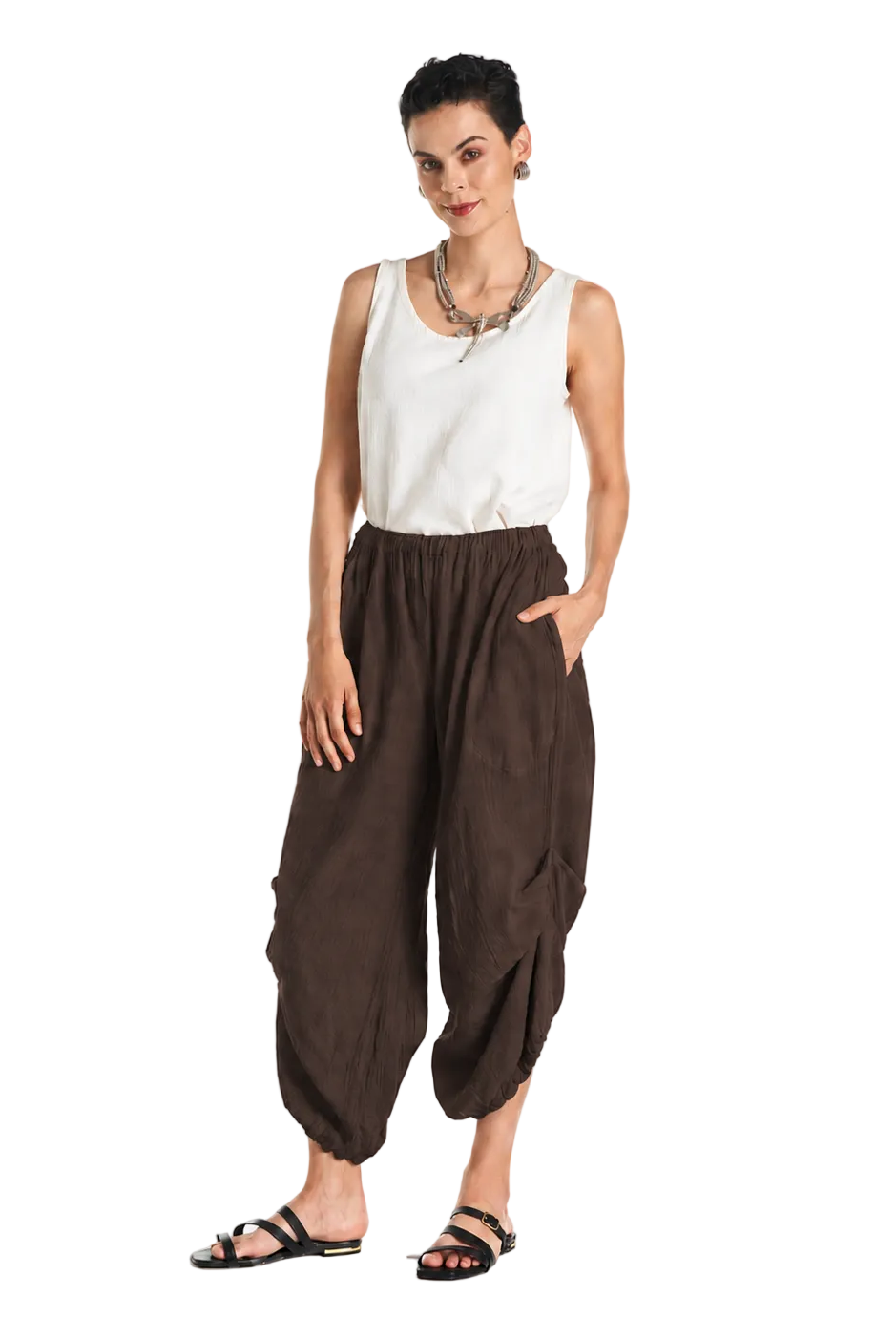 A woman modeling loose brown harem-style pants with a white sleeveless top and black sandals, standing against a plain white studio background.