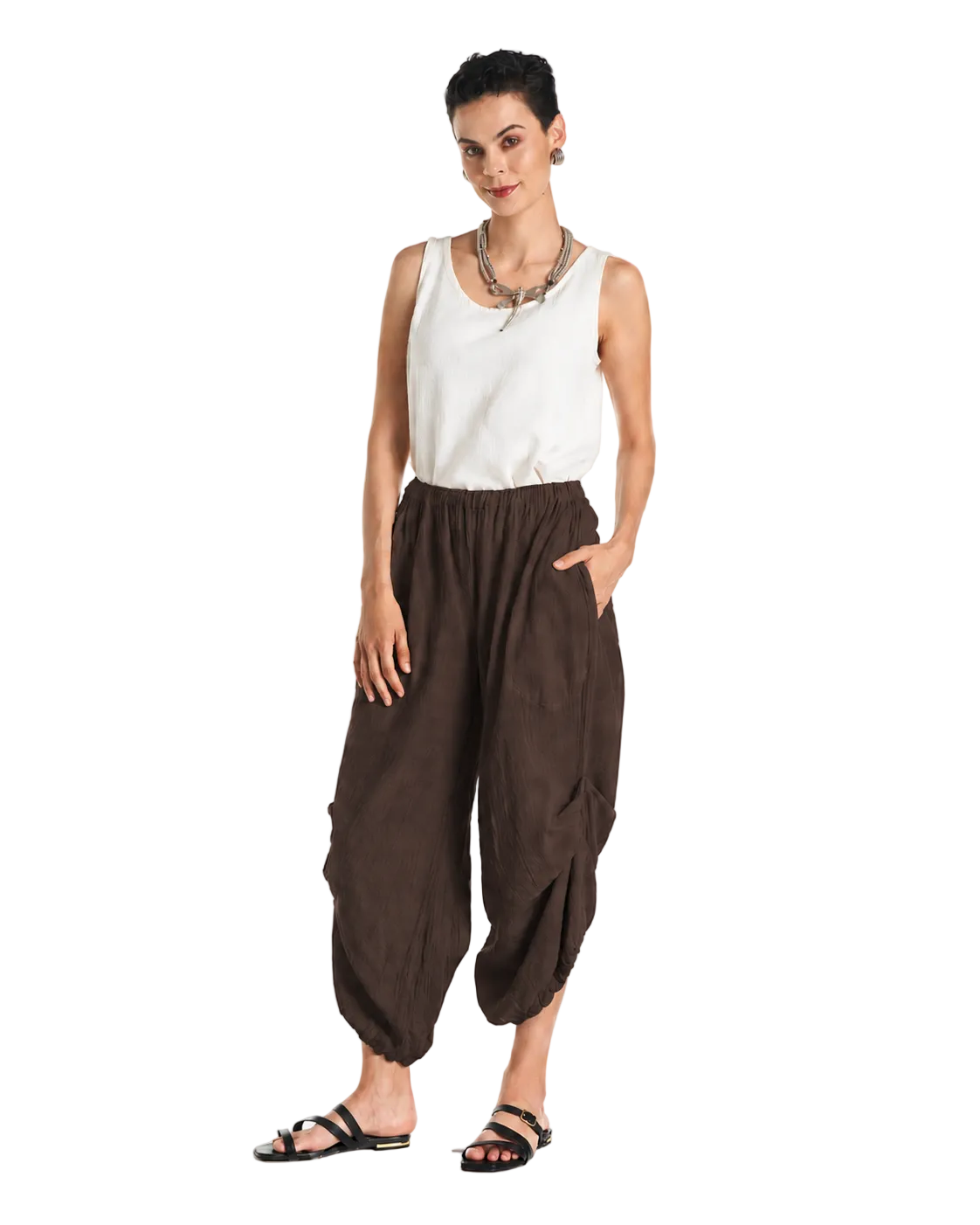A woman modeling loose brown harem-style pants with a white sleeveless top and black sandals, standing against a plain white studio background.