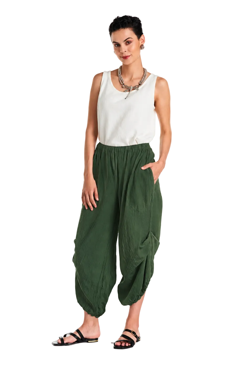 A woman modeling loose, draped olive green harem-style pants with a white sleeveless top and black sandals, standing against a plain white studio background.