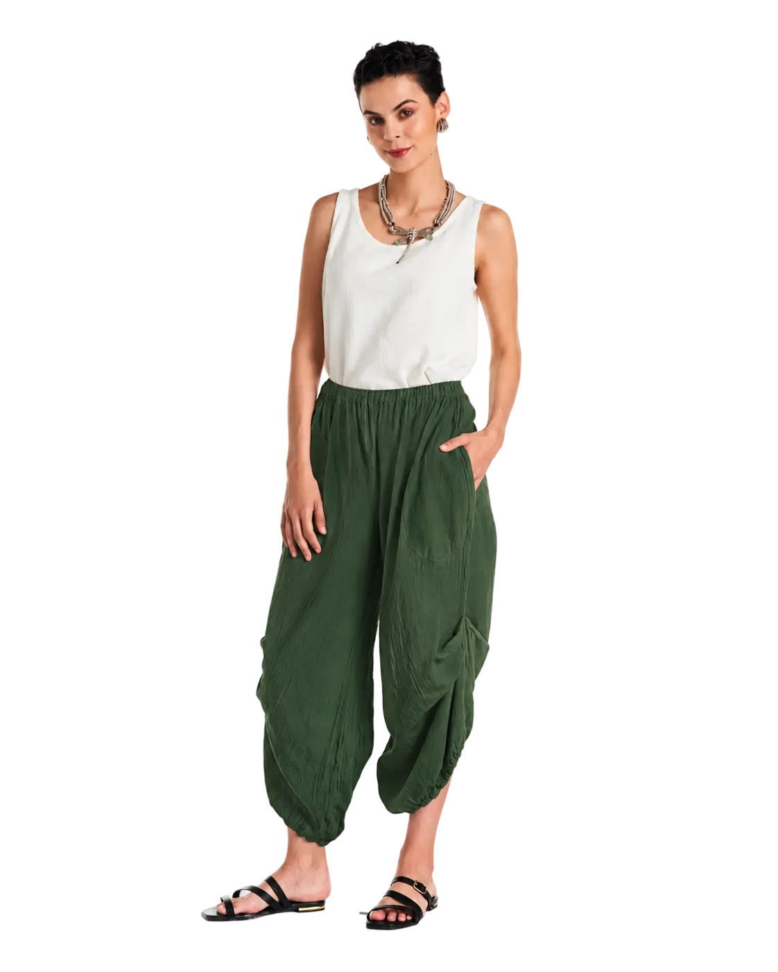 A woman modeling loose, draped olive green harem-style pants with a white sleeveless top and black sandals, standing against a plain white studio background.
