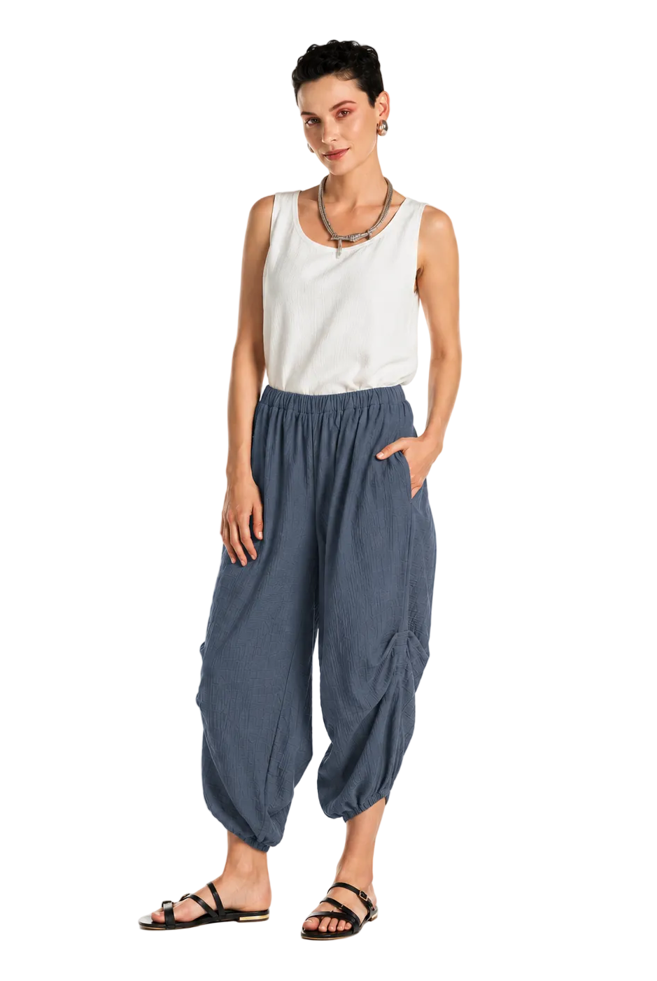 A woman modeling loose, gathered harem-style pants in a muted blue-gray color, paired with a white sleeveless top and black sandals, against a plain white studio background.