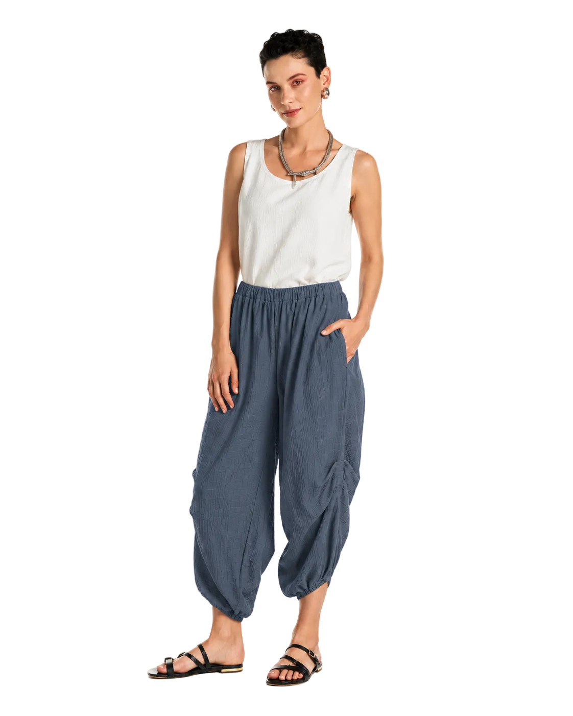 A woman modeling loose, gathered harem-style pants in a muted blue-gray color, paired with a white sleeveless top and black sandals, against a plain white studio background.