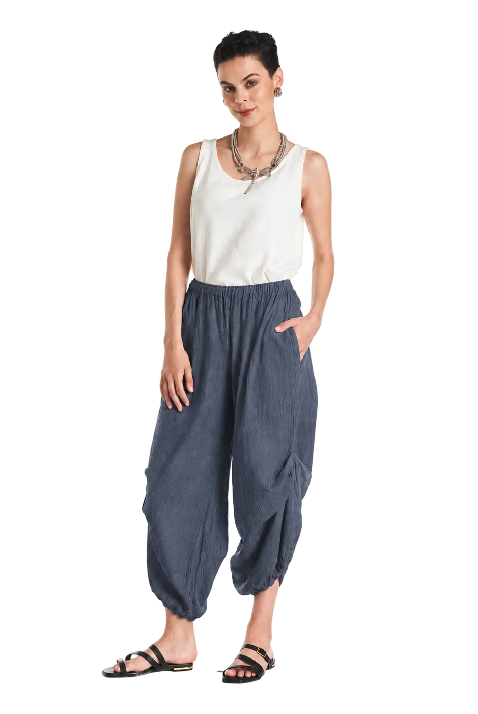 A woman modeling loose, baggy blue pants with a white sleeveless top and black sandals, standing against a plain white studio background.