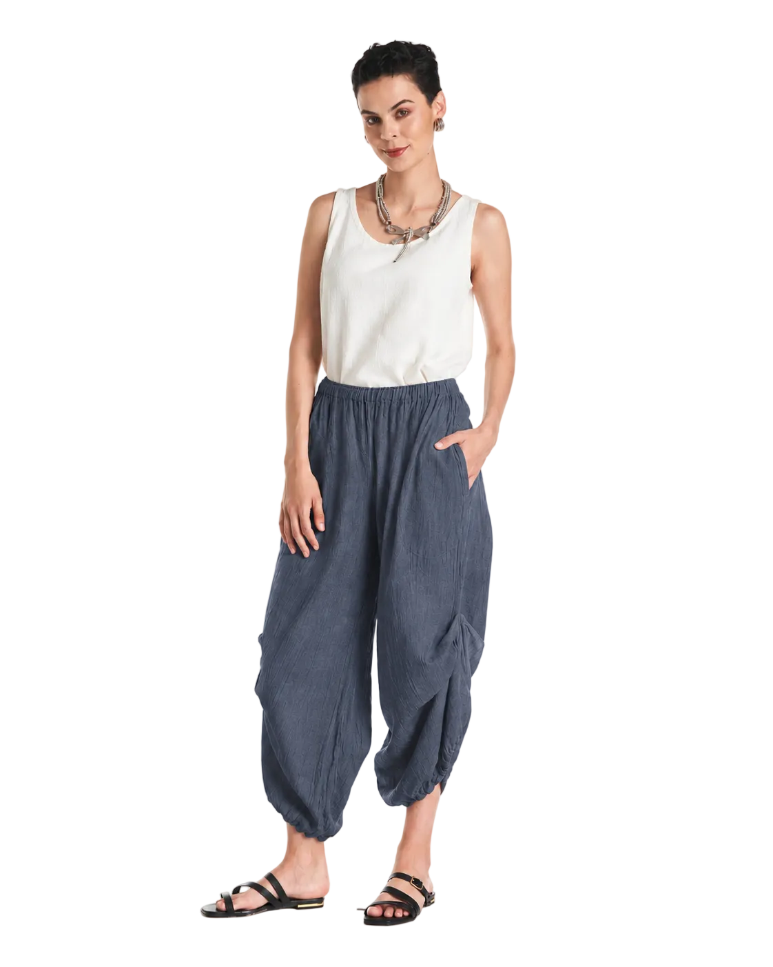 A woman modeling loose, baggy blue pants with a white sleeveless top and black sandals, standing against a plain white studio background.
