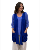 A woman modeling a vibrant royal blue open-front cardigan with pockets over a periwinkle blue top and cream pants, standing in front of a white fabric backdrop.