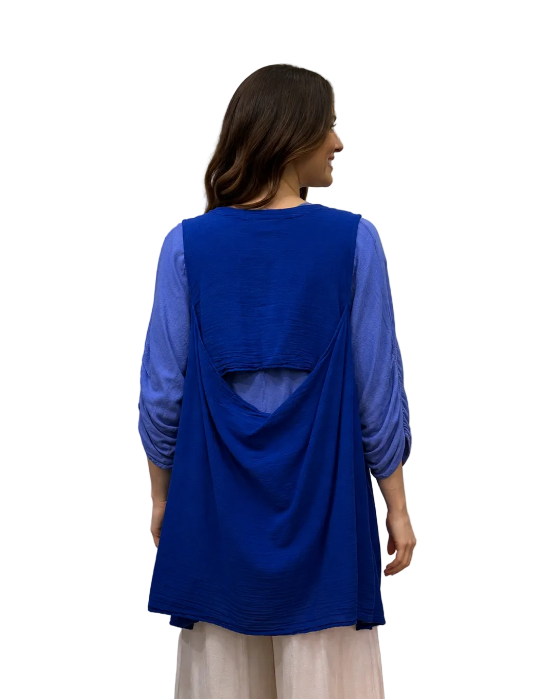 A woman viewed from the back wearing a bright royal blue sleeveless tunic layered over sheer light blue three-quarter sleeves, paired with light cream wide-leg pants, standing in front of a white fabric backdrop.