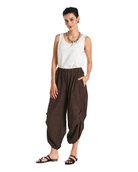 A woman modeling loose brown harem-style pants with a white sleeveless top and black sandals, standing against a plain white studio background.