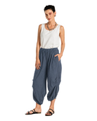 A woman modeling loose, gathered harem-style pants in a muted blue-gray color, paired with a white sleeveless top and black sandals, against a plain white studio background.